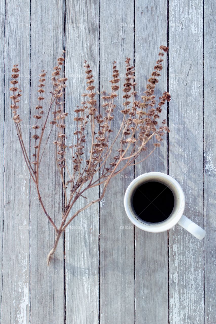 Flat lay of coffee in a white mug next to a dried brown plant on a weathered wooden surface