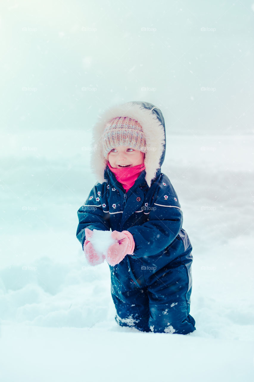 Happy little girl enjoying snow. Child playing outdoors walking through deep snow in wintertime while snow falling. Toddler is wearing dark blue snowsuit