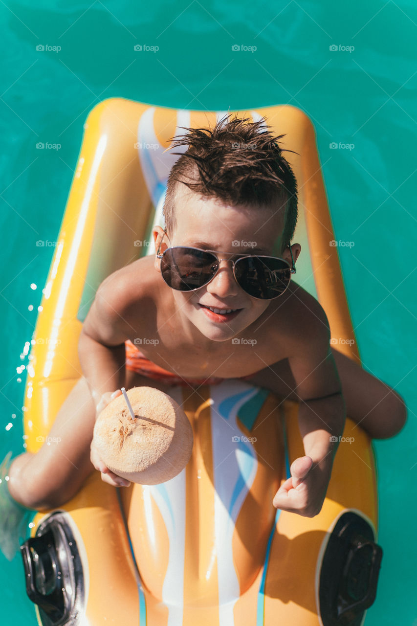 handsome boy of seven years with a coconut in his hands, sits on a swimming mattress, wearing glasses, spends time in the pool, smiles. Summer mood