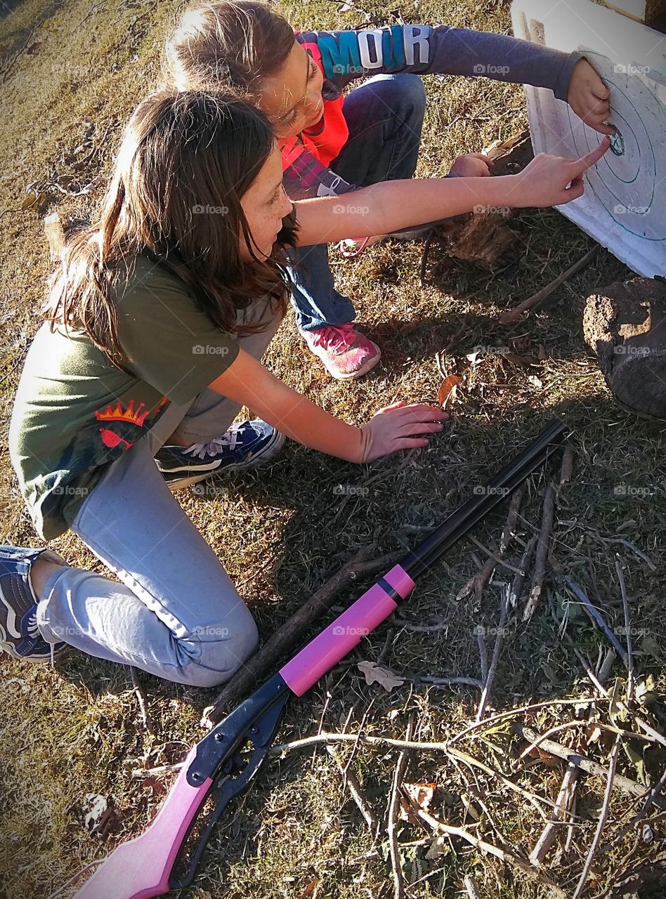 Two cousins, both country girls, younger than 10 years of age - one teaches the other how to shoot her pink Daisy BB gun and conducts target practice.