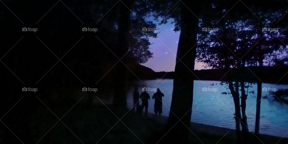 Three men silhouettes stand in the woods overlooking lake water on a beautiful night.