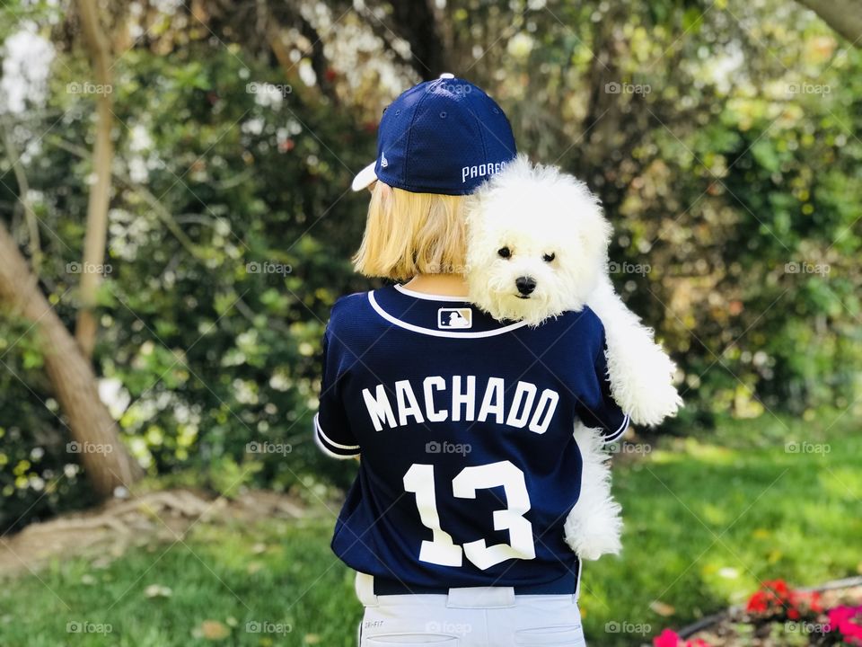 A young blond girl wears a San Diego Padres Machado #13 baseball uniform, and a fluffy white puppy peers over her shoulder. 