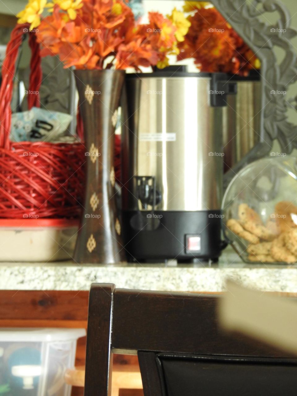 Orange yellow bouquet representing fall autumn behind a woven dark red basket perfect for autumn fall days to warm up with hot chocolate with a large capacity coffee brewer maker and a glass jar with cookies on a kitchen counter and foreground chair.