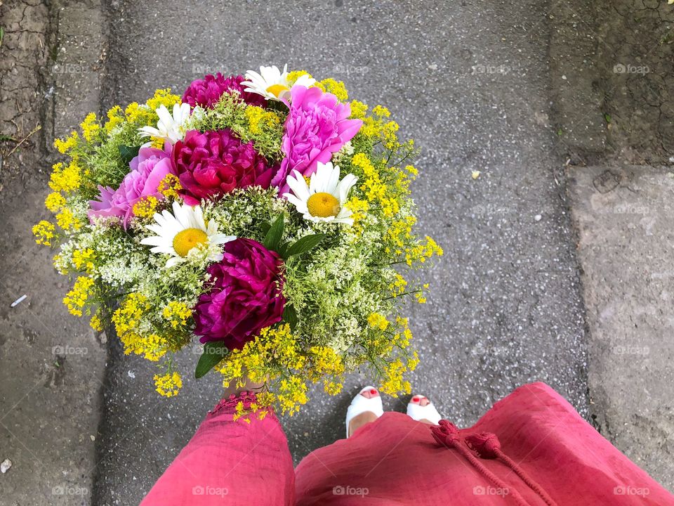 Woman in pink dress holding a giant bouquet of summer flowers consisting of pink peonies, daisies and yellow goldenrod