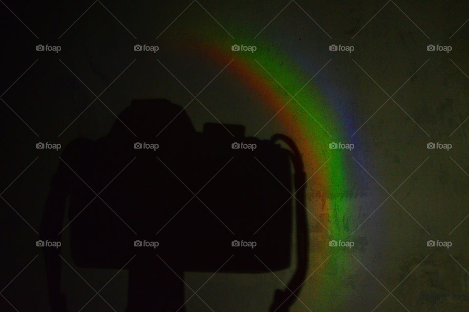 A shadow of a photocamera and a three-colored rainbow on a plain white wall in the dark.