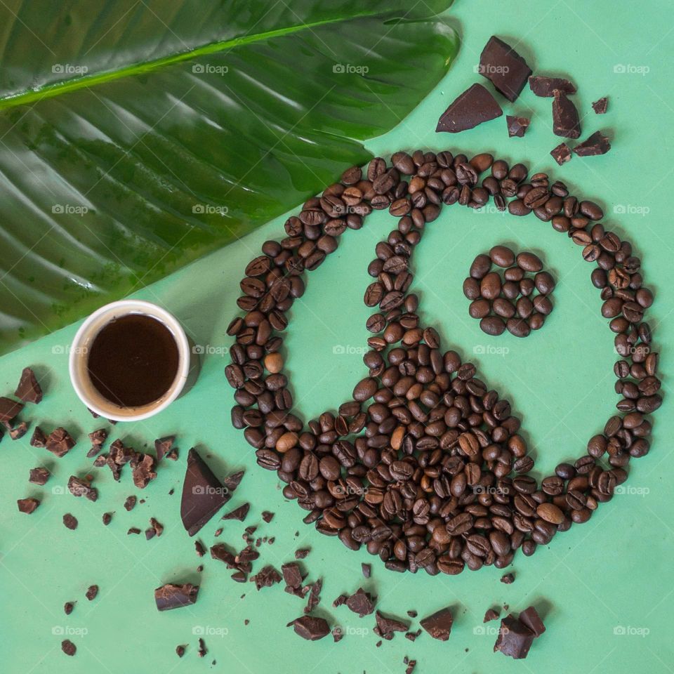 Fairtrade themed flat lay, with coffee beans shaped in the Fairtrade logo, and chocolate and a banana plant leaf against green background 