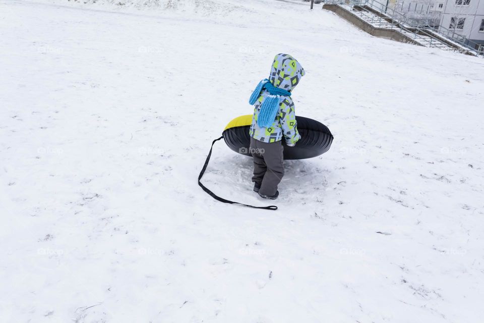 A small, carefree boy walks in the white snow in winter and rides a tubing in the park, near trees in the snow.