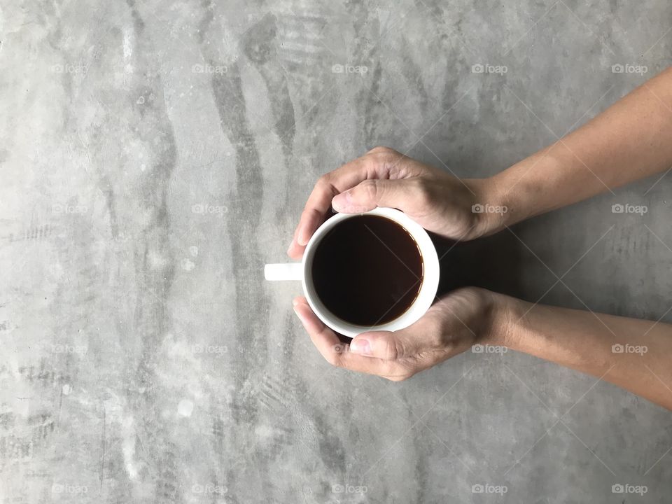 woman holding a white cup of hot black Americano coffee with her both hands on viva board or cement board table in coffee shop with copy space on left side of frame