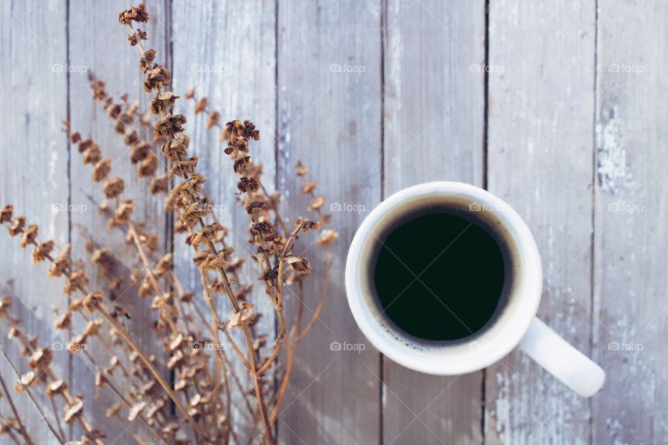 Flat lay of coffee in a white mug next to a dried brown plant on a weathered wooden surface