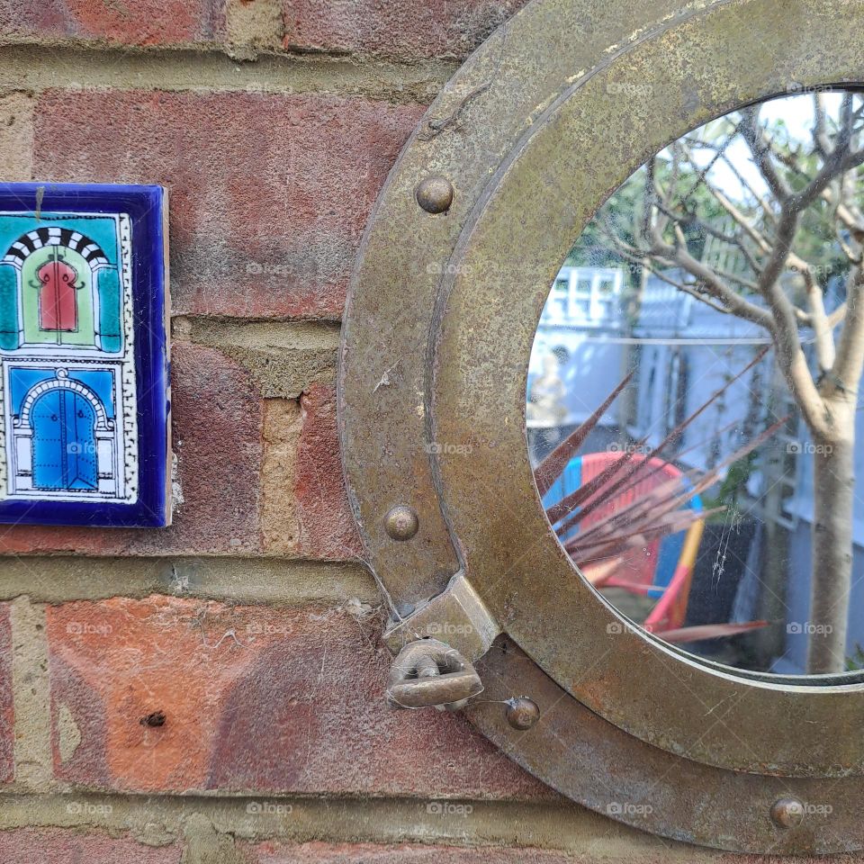 brick wall outside with round brass mirror reflecting a garden fence and chair. u.k