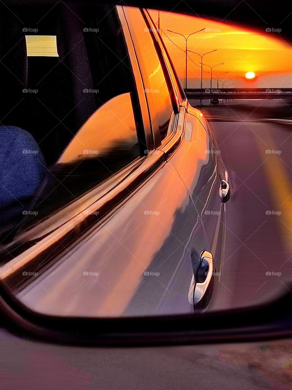 The car is driving on the highway.  The side mirror of the car reflects a blurred road, a bridge and the red sunset sun