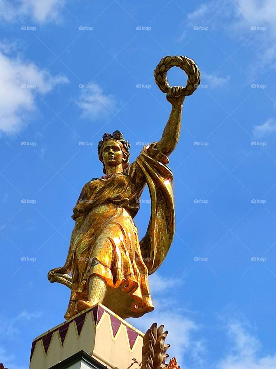 Sculpture of a girl holding out a wreath.  Sculpture in gilded color contrasts with the blue sky and white clouds