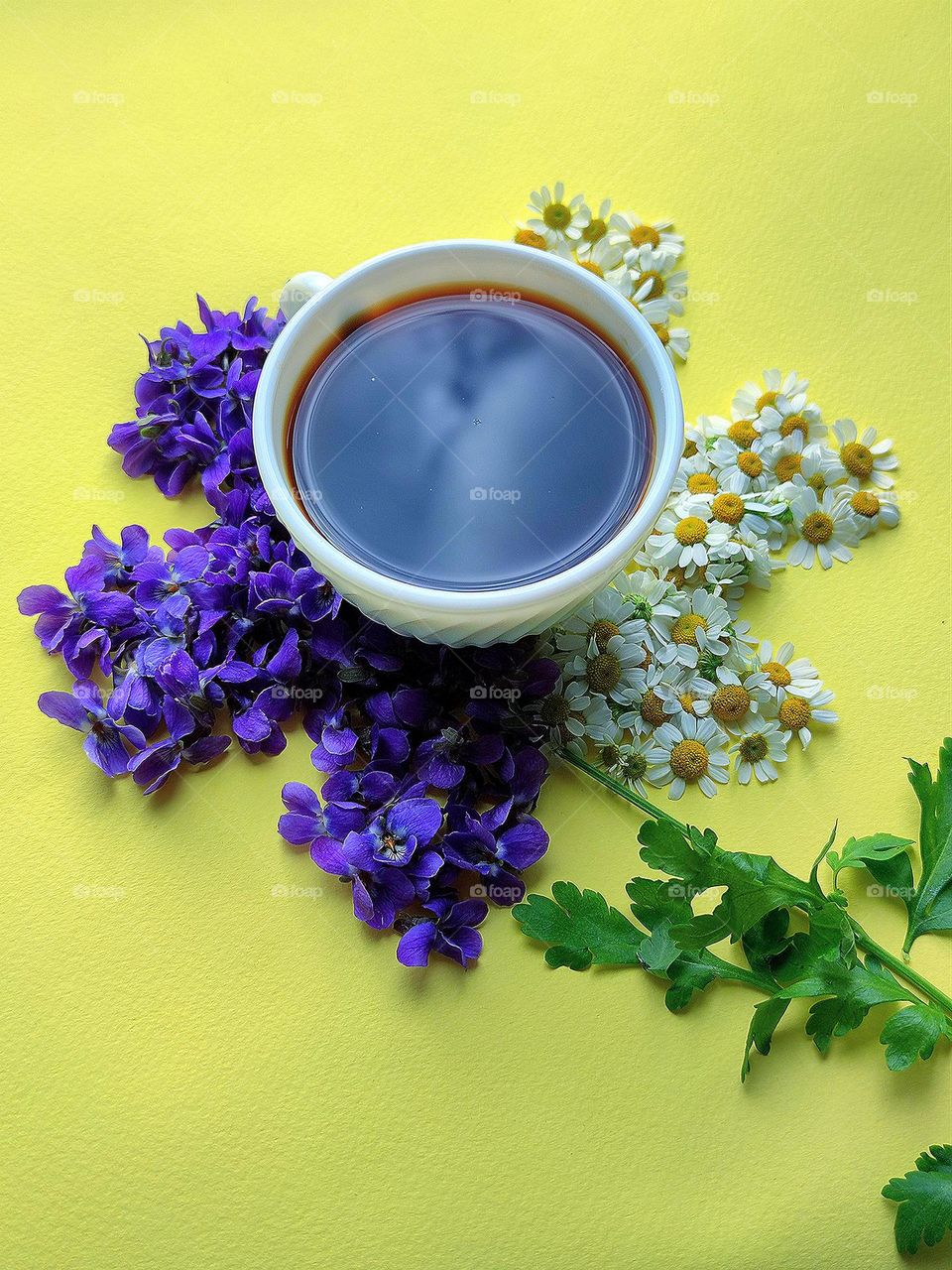 Spring. On a yellow background there is a flower: petals from the flowers of purple violets and white daisies, the core is a white cup of coffee in which the clouds are reflected