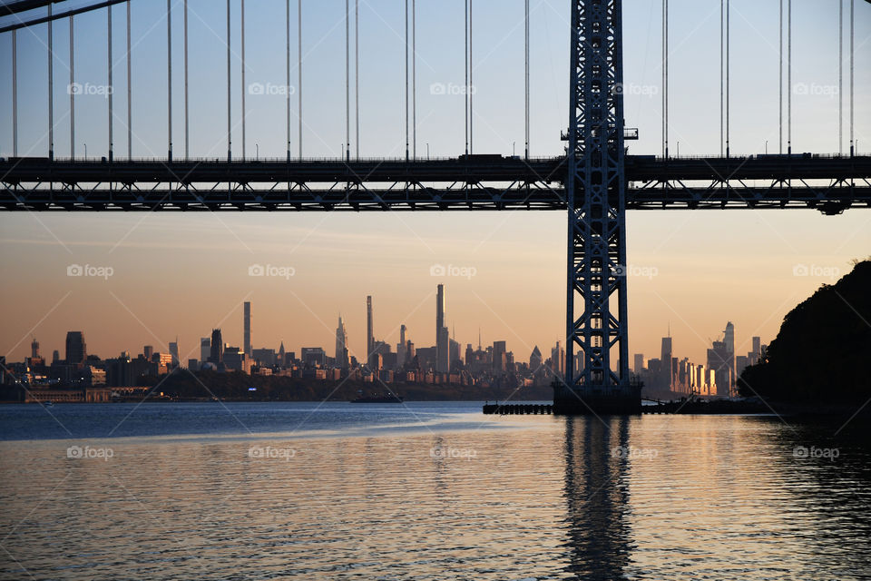 A beautiful sunset highlights the Manhattan cityscape in the far distance off the Hudson river peering through the grandiose presence of the George Washington Bridge.