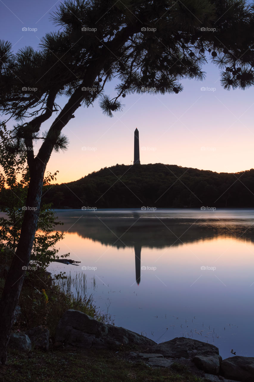 Tall obelisk observation tower on top of a hill reflecting in a body of water. High Point State Park, NJ