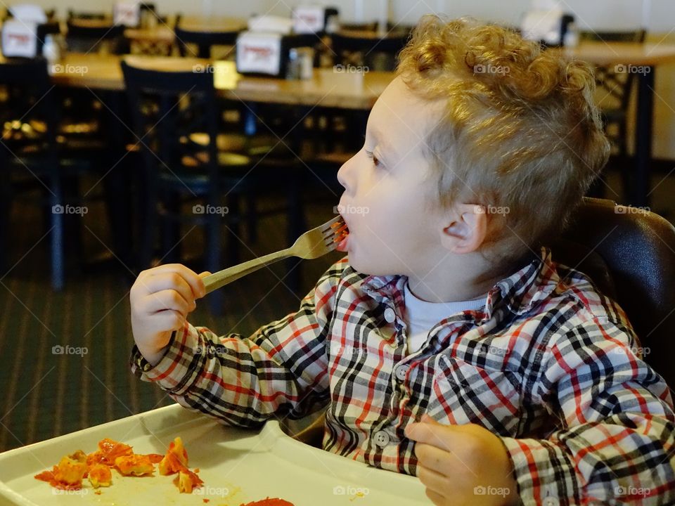 A little boy sitting in a high chair digs into his favorite snack of pepperoni pizza with great vigor. 