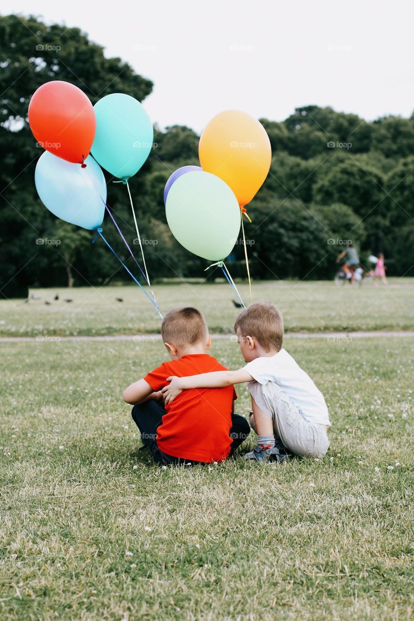 Two little blond boys in a white and red T-shirt sits on a green field, holding a bunch of colorful balloons in his hands, back view 