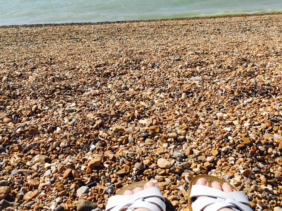 A photo of a rocky beach with a woman’s toes wearing white sandals 
