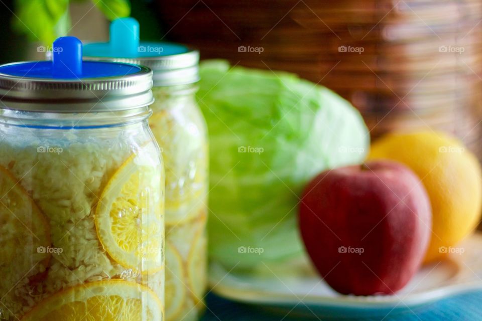 Fermented cabbage, apples and oranges in quart-size mason jars with blue silicone fermenting lids, a cabbage, an apple, and an orange on a white plate on a blue placemat in the background