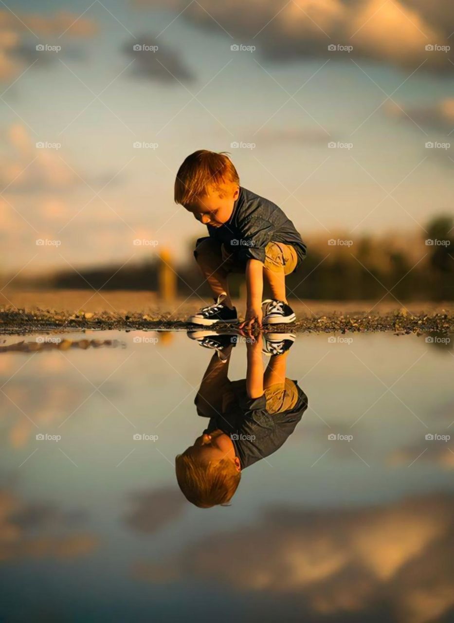 Portrait of a small child crouching and touching the surface of the water that reflects his shadow.