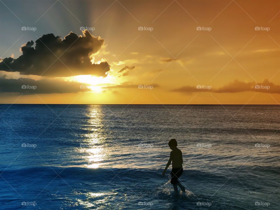 Small boy wading through the blue ocean under the glow of a golden sunset.