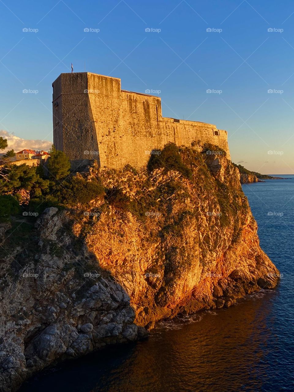 The warm glow of the setting sun against an old medieval fort perched on a cliff on the Adriatic Sea.