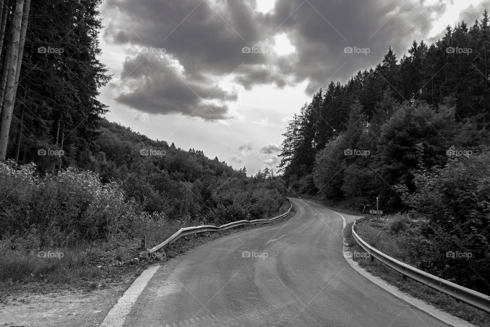 a road through mountains and a forest with a cloudy sky