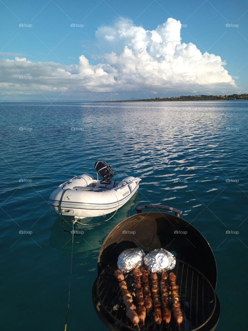 View of dinghy or R.H.I.B / Rib and Barbecue off the back of a Yacht, looking out to huge Clouds in the distance and seen across the Seas calm surface in the reflections on the Oceans Blue and Green Waters.