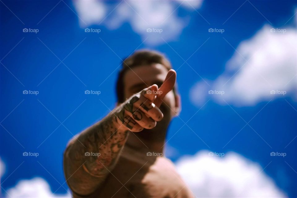 Tattooed man pointing to the camera, with hand and arms tattoos. A beautiful blue sky is on the background.