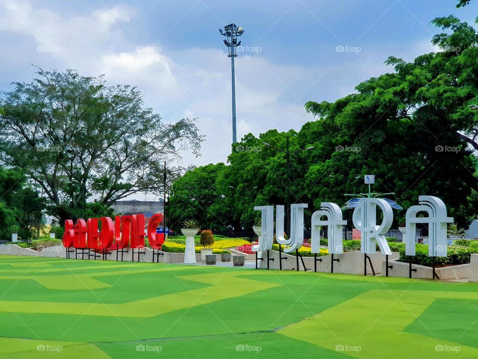 Bandung city center park, Alun Alun Bandung city, light green artificial grass, tall shady green leaf tress, sky refrects blue, wahite clouds, stretches the words "Bandung Juara = Bandung Champion" red and white