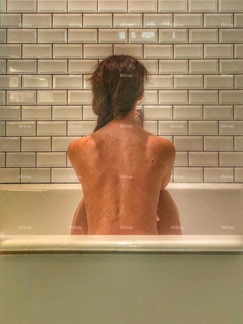 Young woman sits naked in bath with her back to the camera with metro tiles in background
