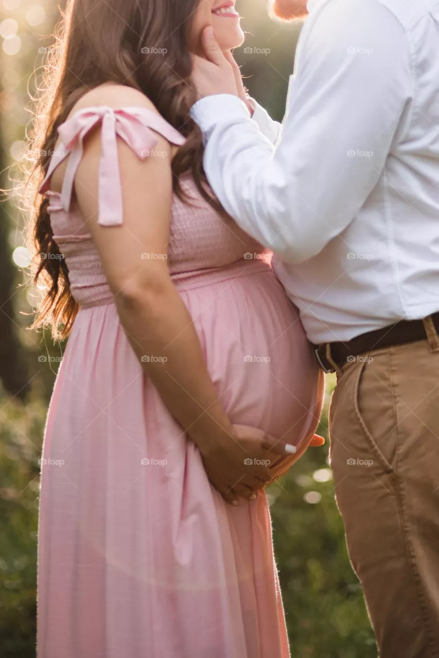 Close up of a beautiful pregnant couple, showing love to one another with a kiss in an outdoor setting. Focus is on the fully grown baby bump, as the warm emotions  of awaiting baby’s arrival take over.