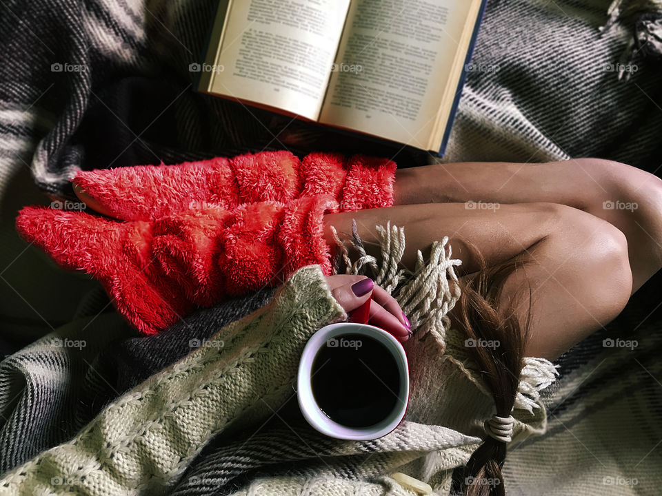 Overhead view of a young woman wearing pink home boots and enjoying a cup of coffee with a book in a cozy warm bed