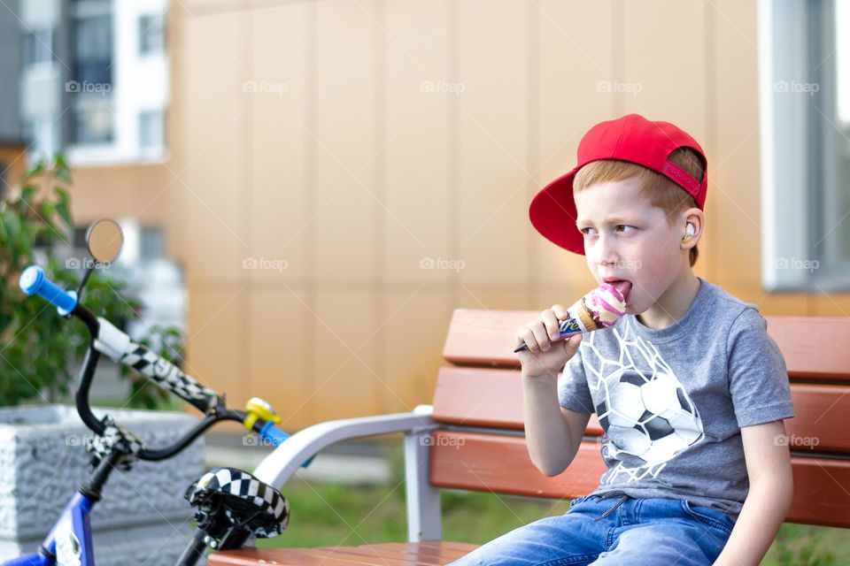 A child in a red cap on the street sits on a bench eats ice cream next to a bicycle summer