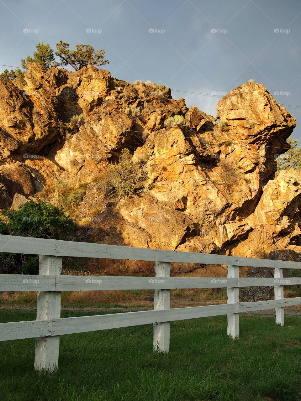 A golden glow covers the beautiful and rugged geology in rural Central Oregon after a rainstorm with a white fence in the foreground. 