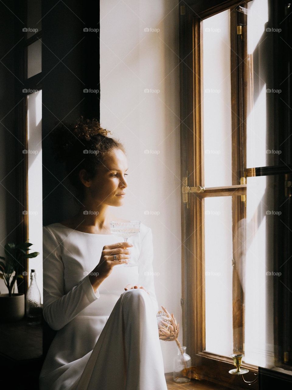 Young beautiful woman with curly hair wearing white dress sits on the window and drinks champagne, portrait of woman, looking at window, lifestyle, at home, relax.