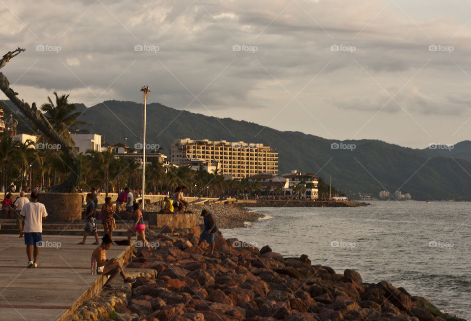 Partially seen in the foreground is one of the many bronze statues that dots the length of the Malecon, while in the background swimmers swim and the Voladores of Papantla flying men begin their traditional performance at the top of the white pole from which they swing.