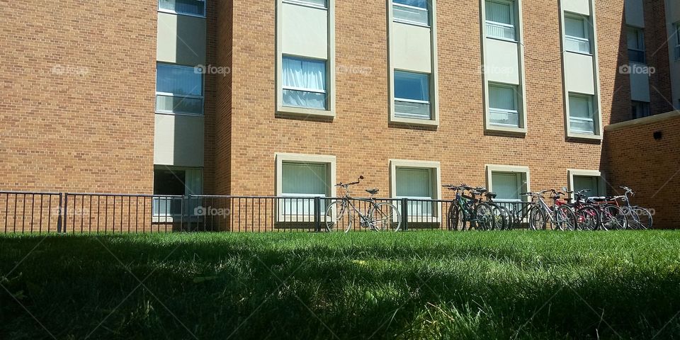 several bikes on a rack outside of a brick dorm building on a sunny day