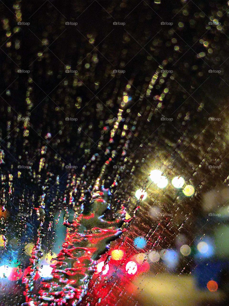 Water splashes on the windshield of a car.  One can see the colorful lights of the big city