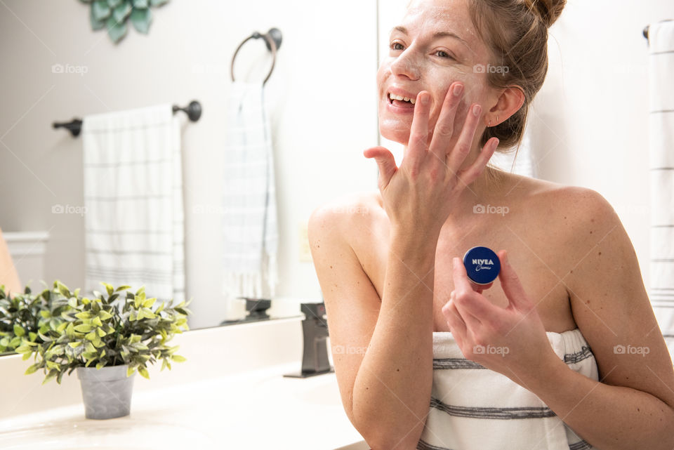 Portrait of a smiling young millennial woman applying Nivea cream to her face in a bathroom