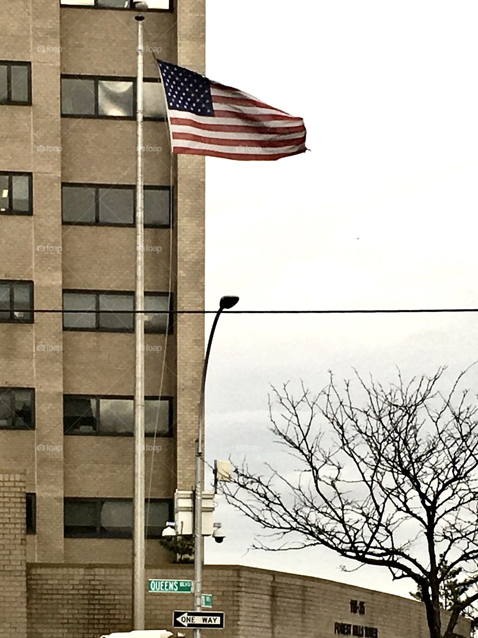 Flag, American flag, flying, high, flag pole, city, street, New York, street sign, one way, bare tree, tree, outside, exterior, security camera, light pole, power line, electric line, building, windows, Queens sky, red, white, blue, stars, stripes