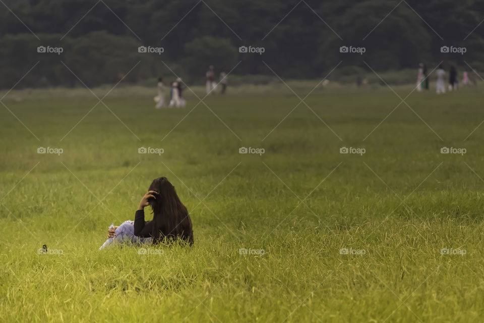Pretty, cheerful girl sitting on green grass and rests. A young woman or girl on green meadow watching and enjoying nature summer evening. Holiday event travel vacation concept ( Kolkata India )