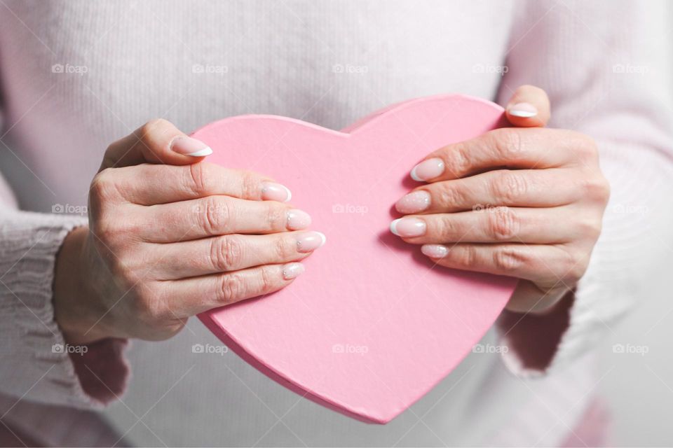 Hands of a young Caucasian girl with a beautiful delicate classic manicure holding a pink cardboard heart-shaped box standing in the room,side view. Concept beautiful woman's manicure,valentine's day,love day.