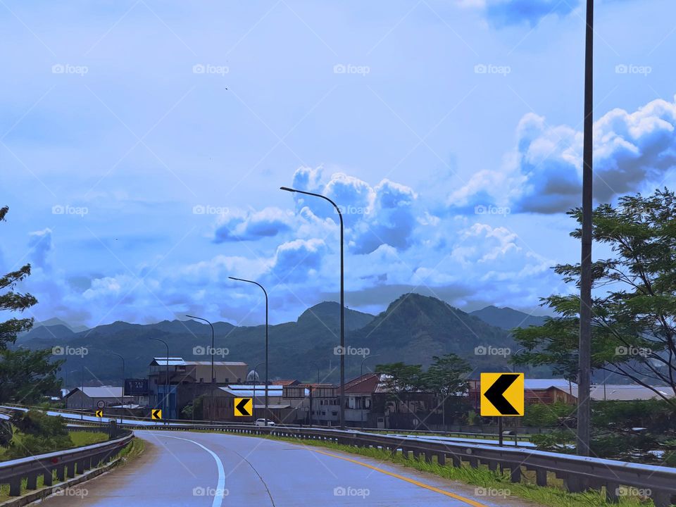 Highway traffic sign, chevron sign to the left, base color for yellow sign, black chevron sign, view of driving on the highway, the sky refracts blue, and the clouds are bright white