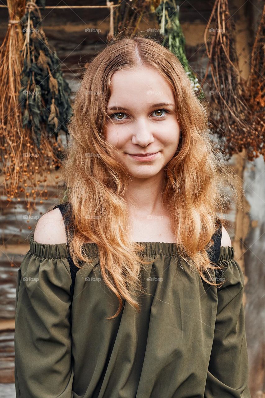 Portrait of young woman standing in front of old cottage and hung some withered species of herbs. Girl is wearing green dress, looking at camera