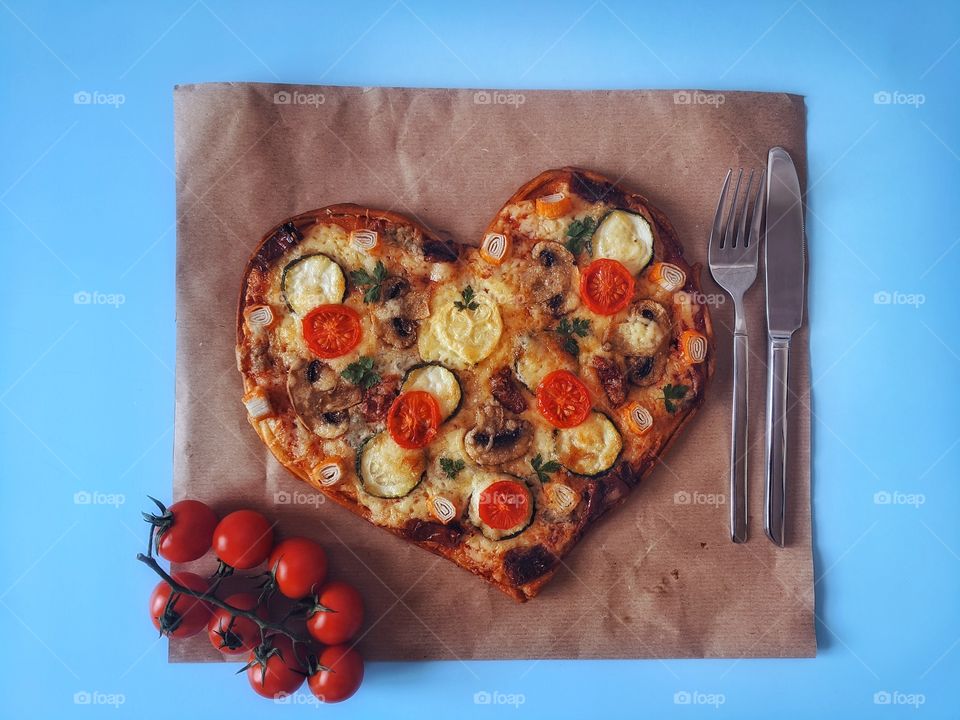 Delicious vegetarian pizza in the shape of a heart for Valentine's Day with tomatoes, vegetables and cheese on a blue background.