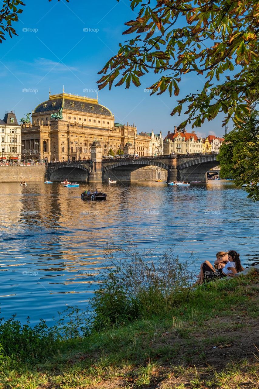 A group of young women sitting on the banks of the Vltava River in the city of Prague gaze attentively at the buildings on the opposite shore