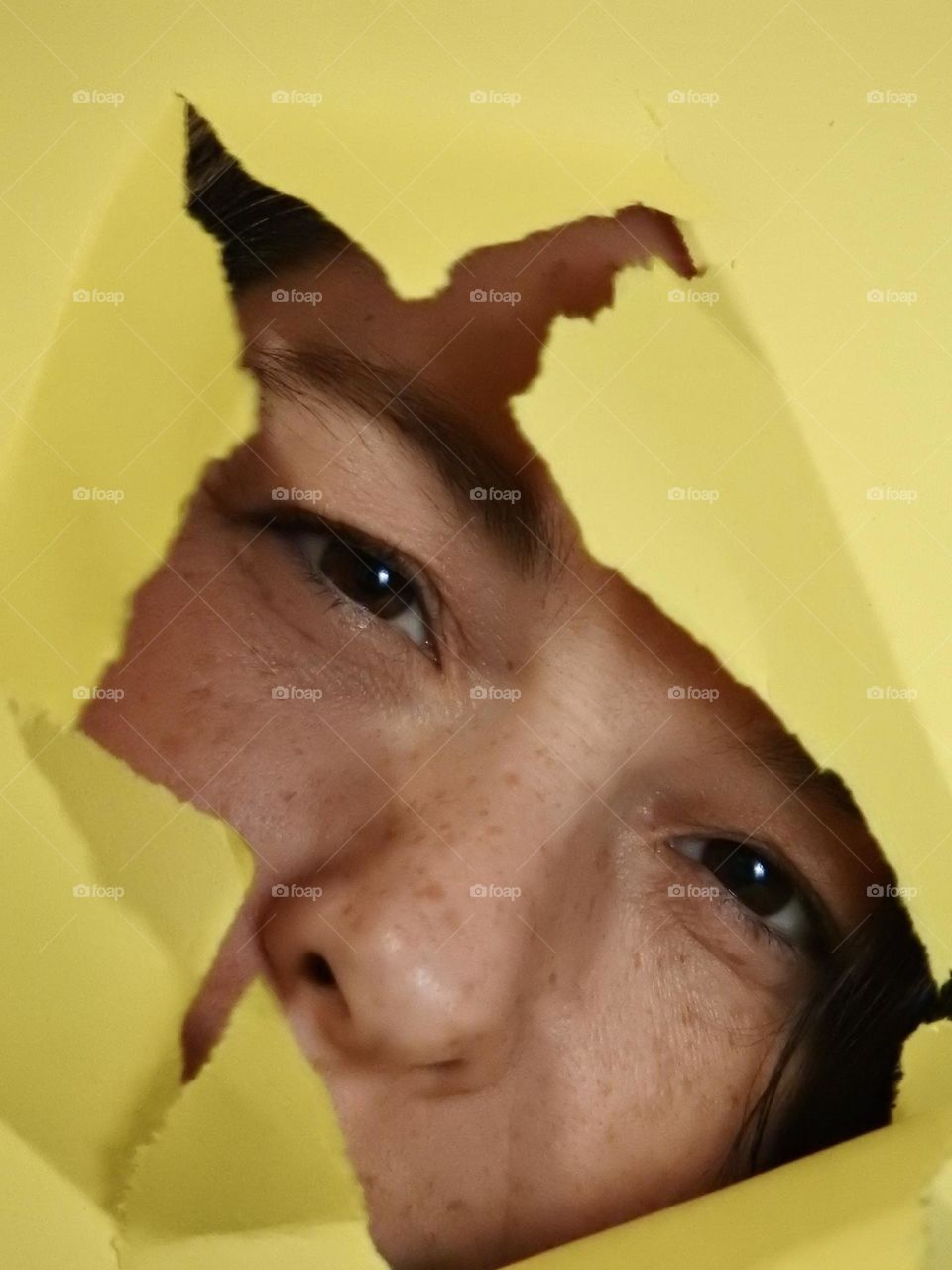 Face skin in the forties of a woman with freckles and brown hair seen through a broken yellow paper.