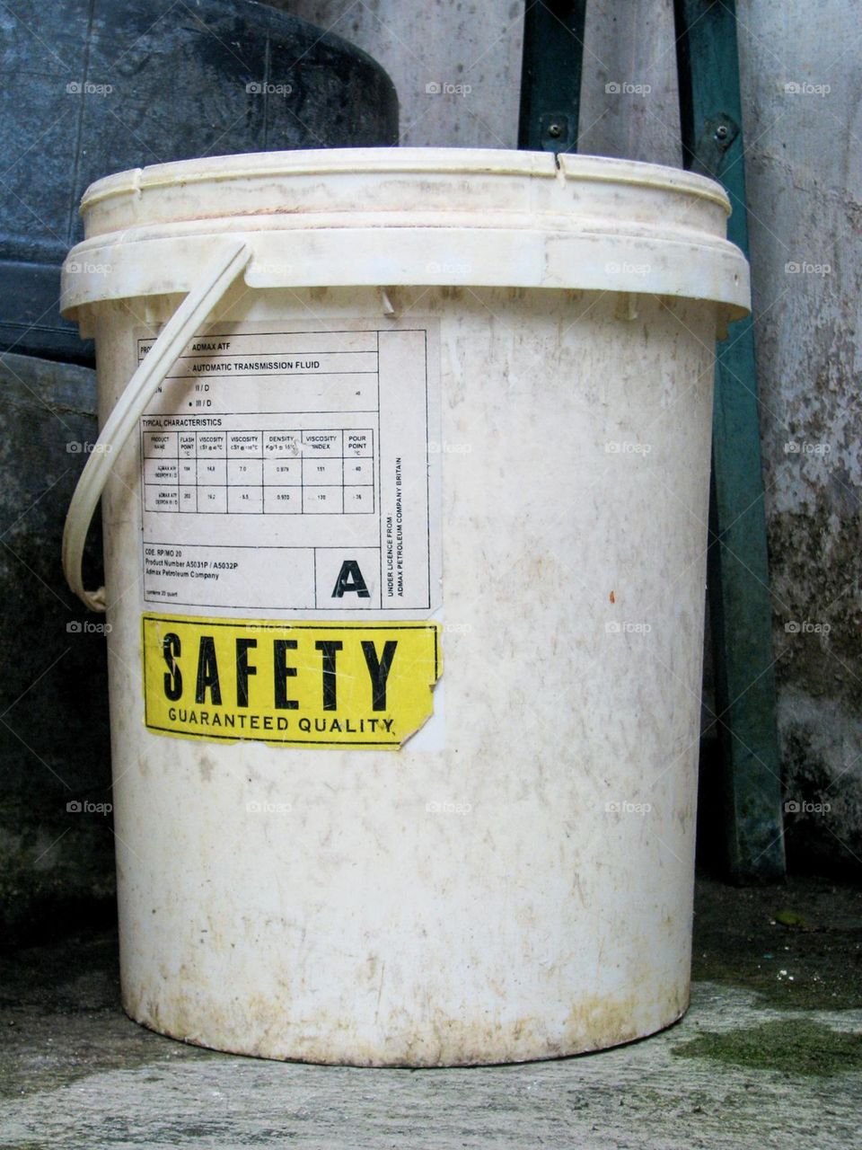 Close-up of a worn and dirty white bucket, with a label and some text on it, placed near a concrete wall