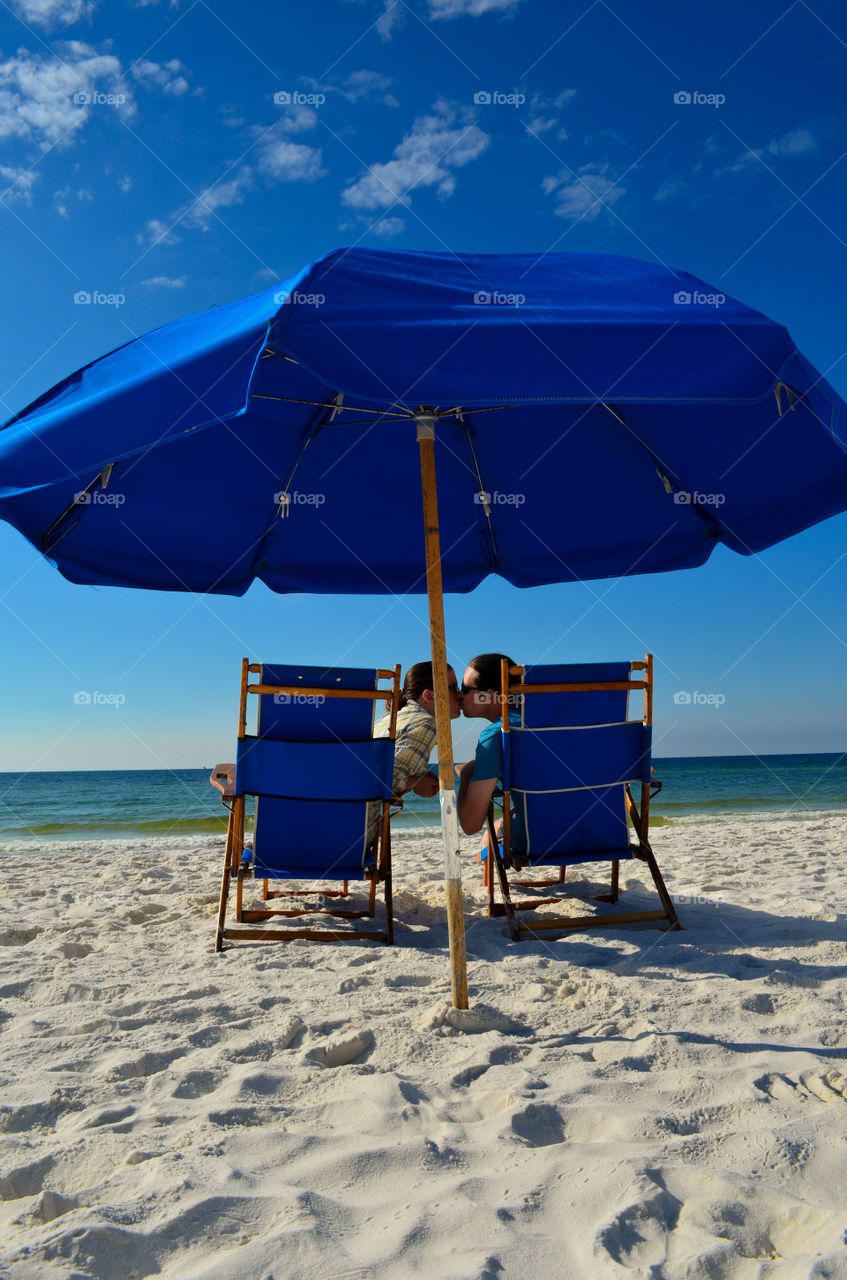 Couple kissing in blue beach chairs under an umbrella at the beach